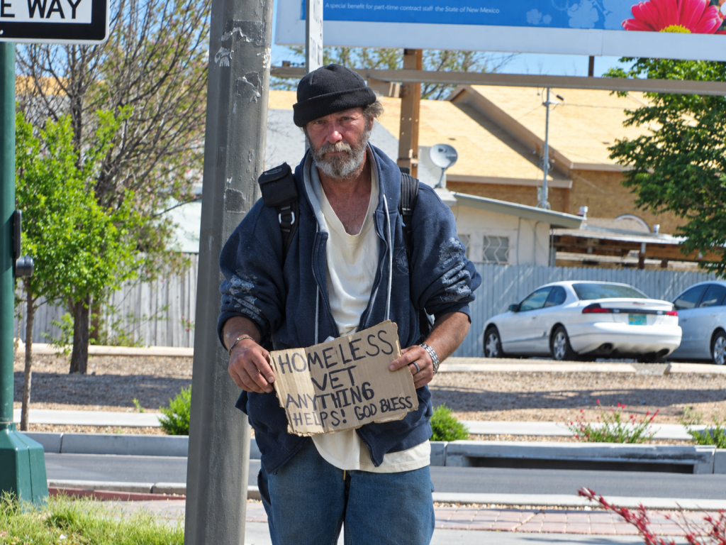 A man standing on the sidewalk holding a sign that reads 'HOMLESS VET ANYTHING HELPS! GOD BLESS'. He wears a blue jacket and has a backpack, with trees and parked cars in the background.