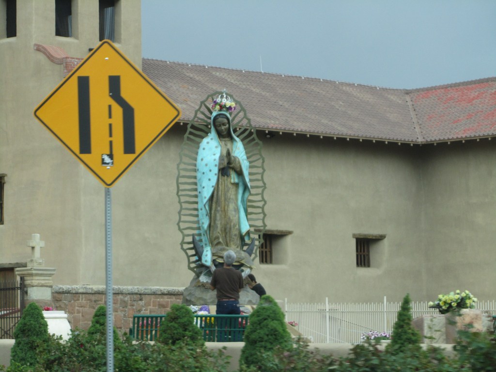 A statue of the Virgin Mary dressed in blue and white, surrounded by greenery, with a person in a brown shirt standing in front of it, and a warning traffic sign indicating a road bend in the foreground.