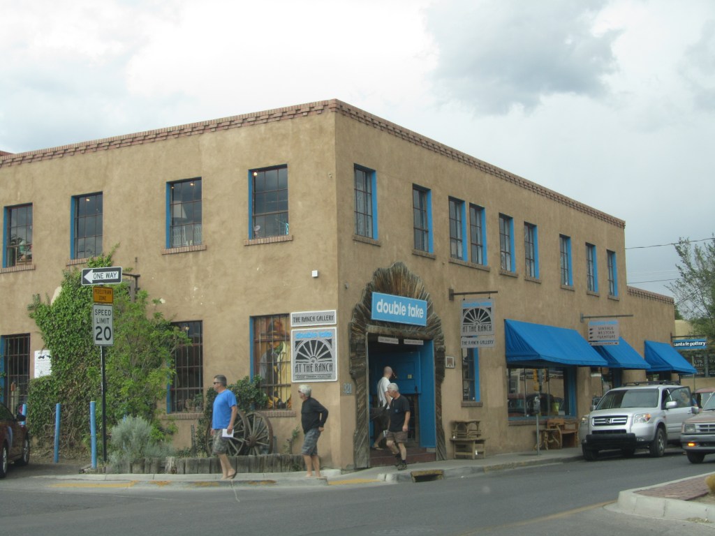 Exterior view of a two-story adobe-style building featuring shops, including 'double take' and 'the ranch gallery,' with pedestrians walking in front and blue awnings over windows.