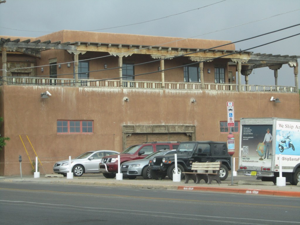 A brown adobe building with a wooden balcony and several parked cars in front, including a silver sedan and a black jeep.