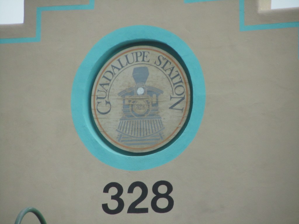A circular sign featuring the text 'Guadalupe Station' and an illustration of a steam locomotive, set against a beige wall with a turquoise border, alongside the number '328'.
