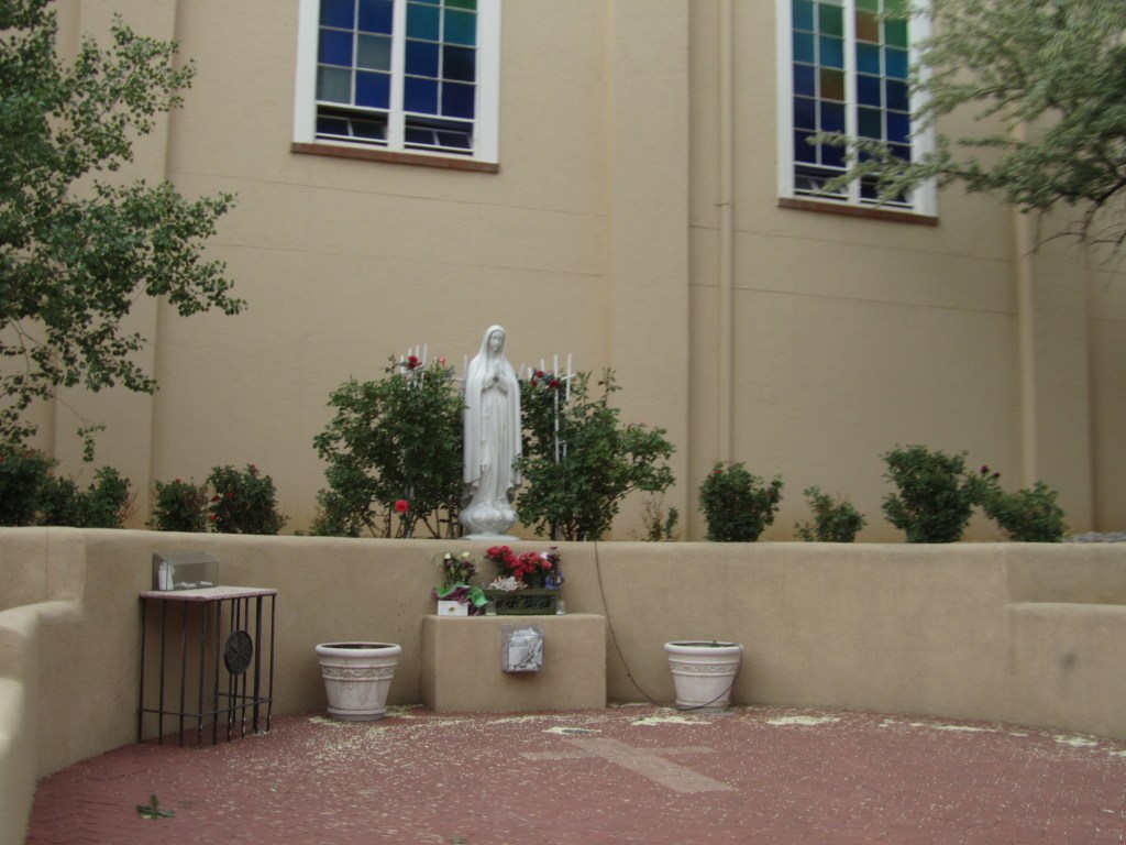 A serene outdoor religious area featuring a statue of a woman, surrounded by flowers and greenery, with a building in the background and colorful stained glass windows.