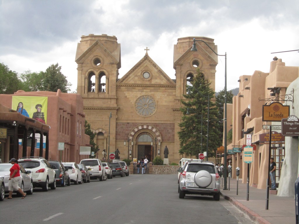 Street view of a historical church with twin bell towers, surrounded by adobe buildings and parked cars.
