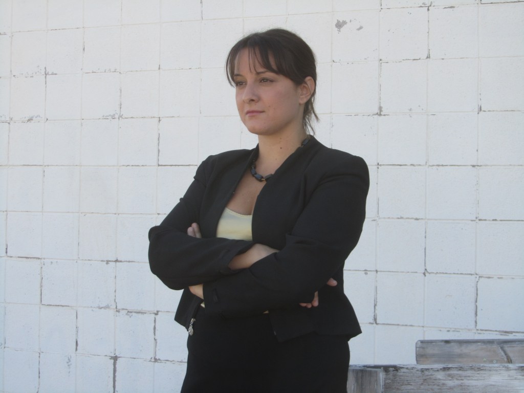 A woman standing confidently with her arms crossed, wearing a black blazer and a yellow top against a white brick wall.