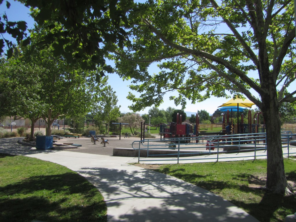 A view of a playground surrounded by trees, featuring play equipment and a pathway leading into the area on a sunny day.