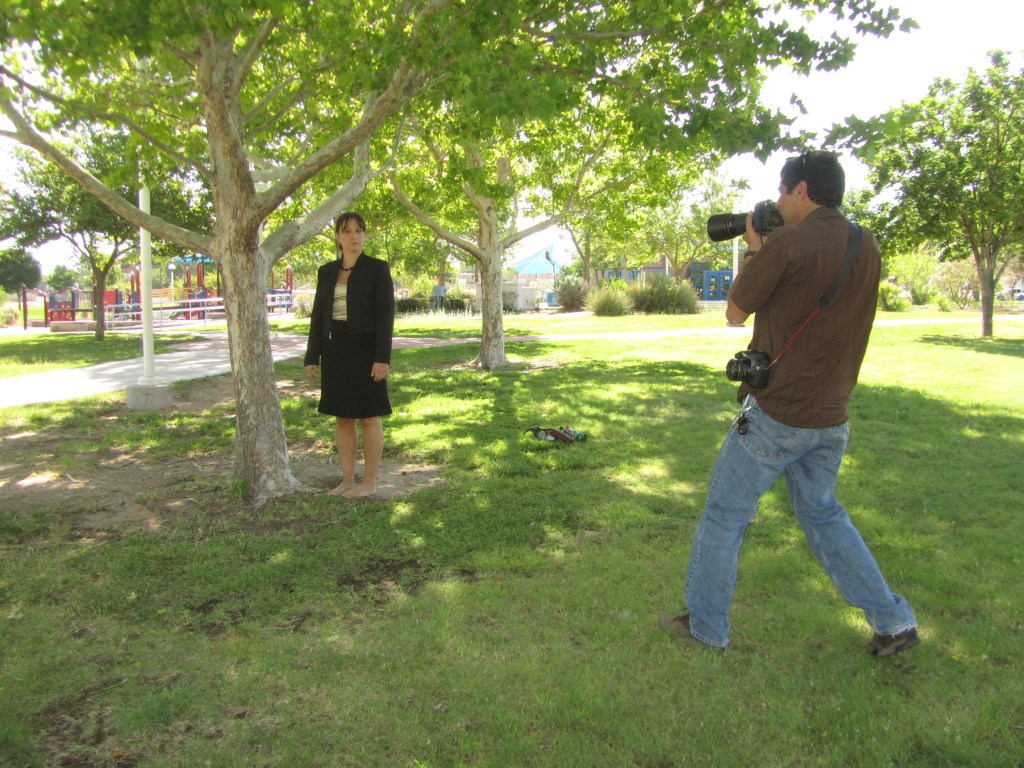 A woman in a black suit stands barefoot in a park, while a man with a camera captures her image. Lush green trees and a playground are visible in the background.