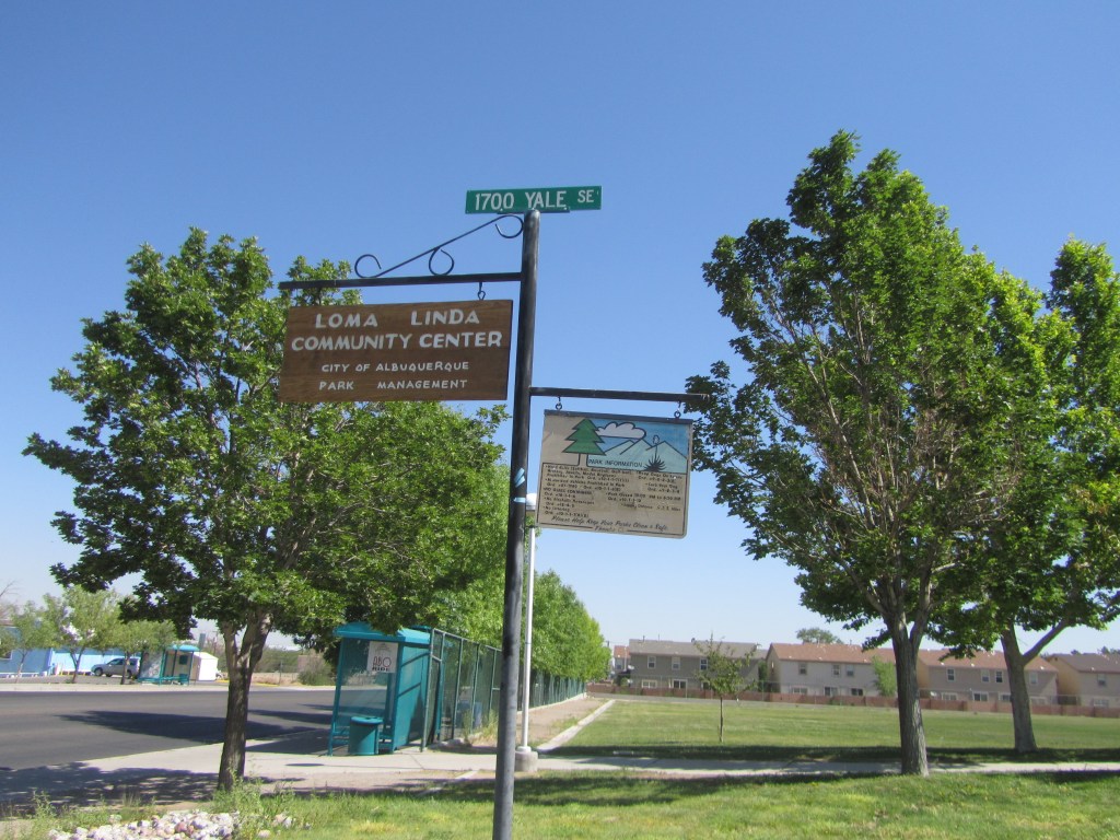 Sign for Loma Linda Community Center with street sign '1700 Yale SE' and park information sign, surrounded by green trees and a clear blue sky.