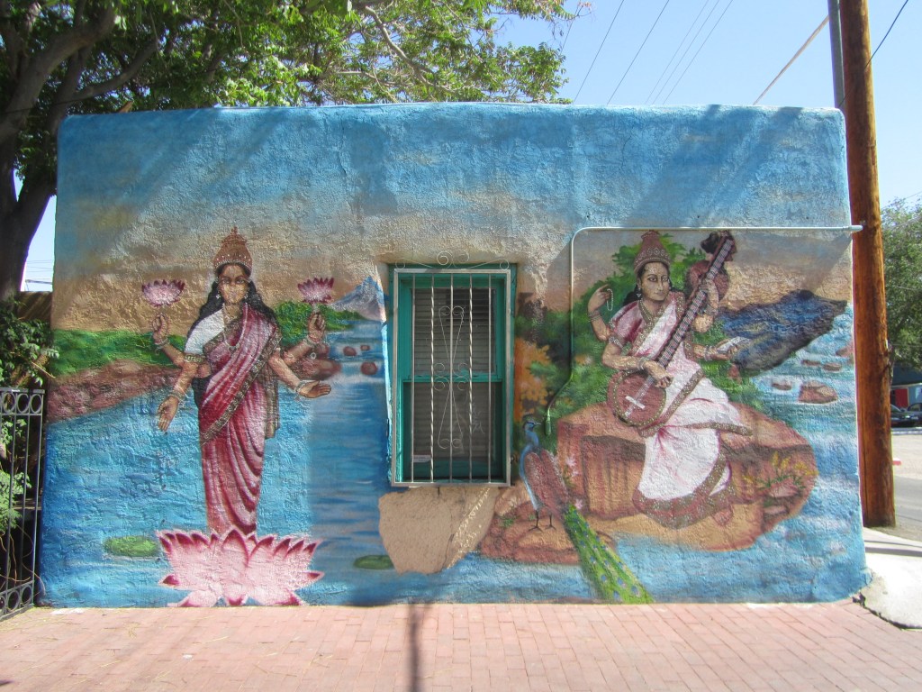 A colorful mural depicting two traditional Indian goddesses, one holding lotus flowers by a river and the other playing a musical instrument, set on a blue wall.