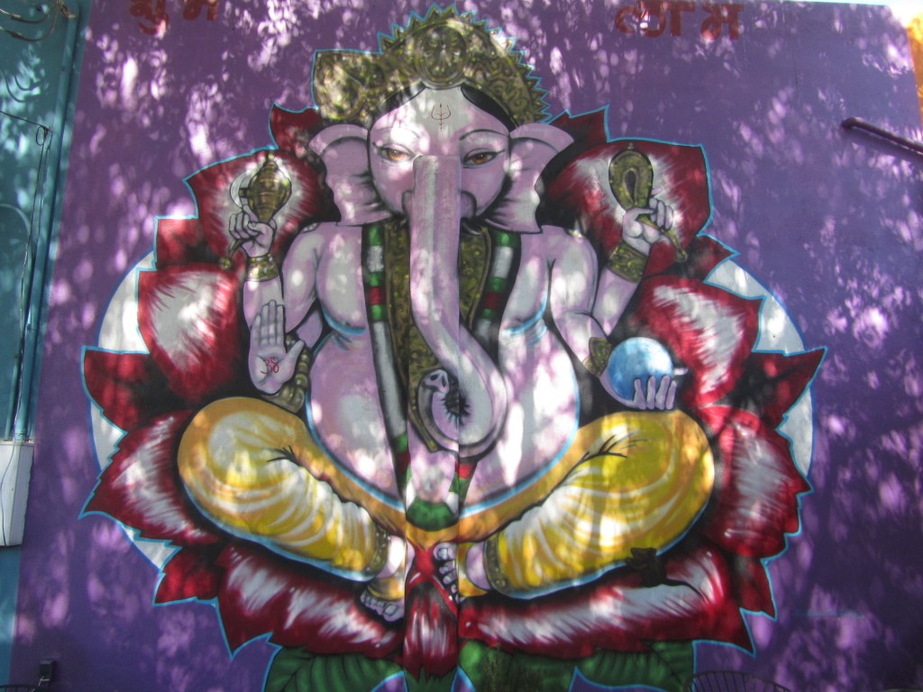 A colorful mural depicting the Hindu deity Ganesha, seated in a lotus position. Ganesha has an elephant head and multiple arms holding symbolic items, surrounded by vibrant floral designs.