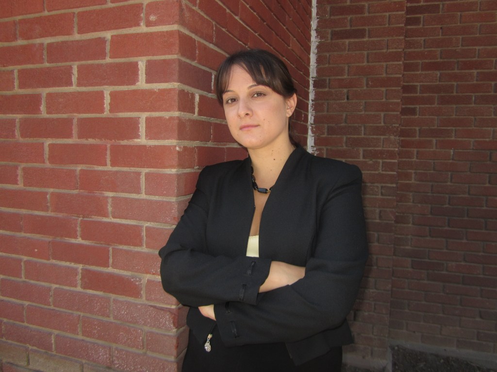 A woman standing confidently with her arms crossed, wearing a black blazer, in front of a brick wall.