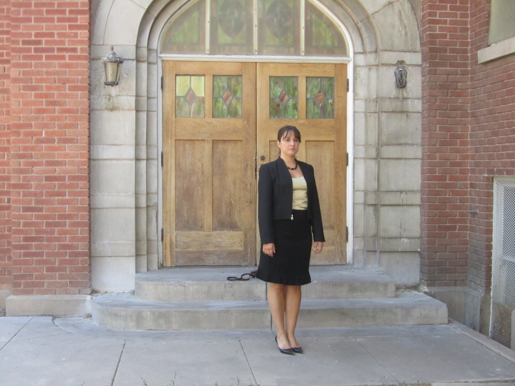 A woman in professional attire stands on the steps of a building with wooden double doors, surrounded by a brick facade.