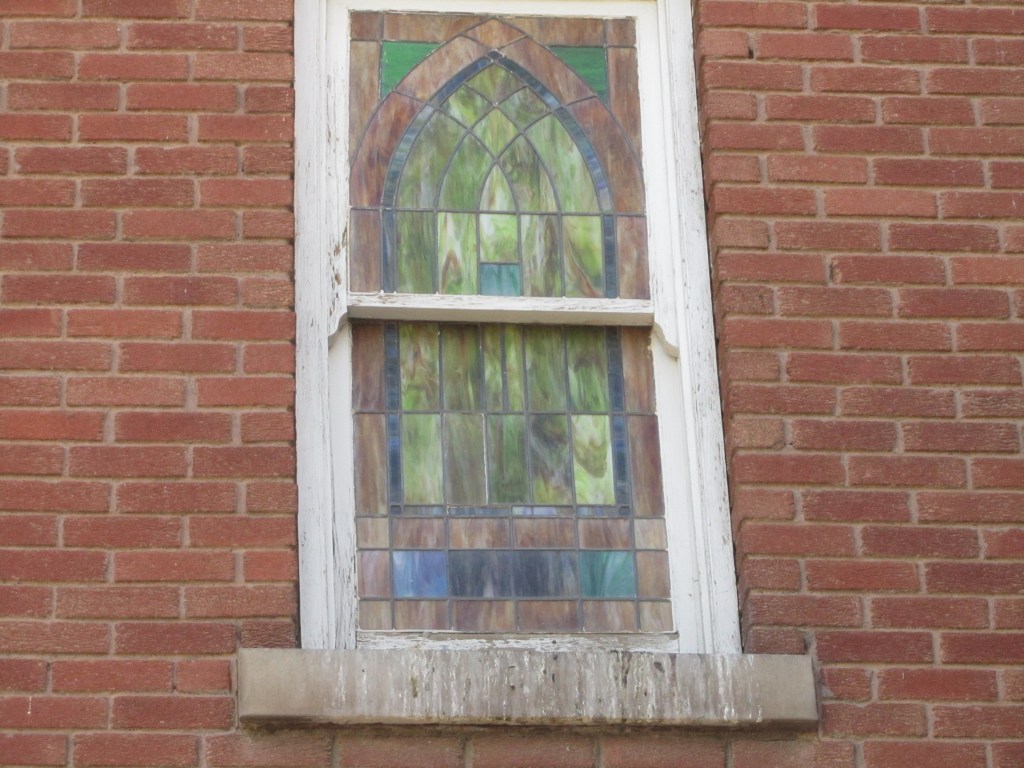 Stained glass window with an arched design, set in a brick wall, featuring colorful glass panels.
